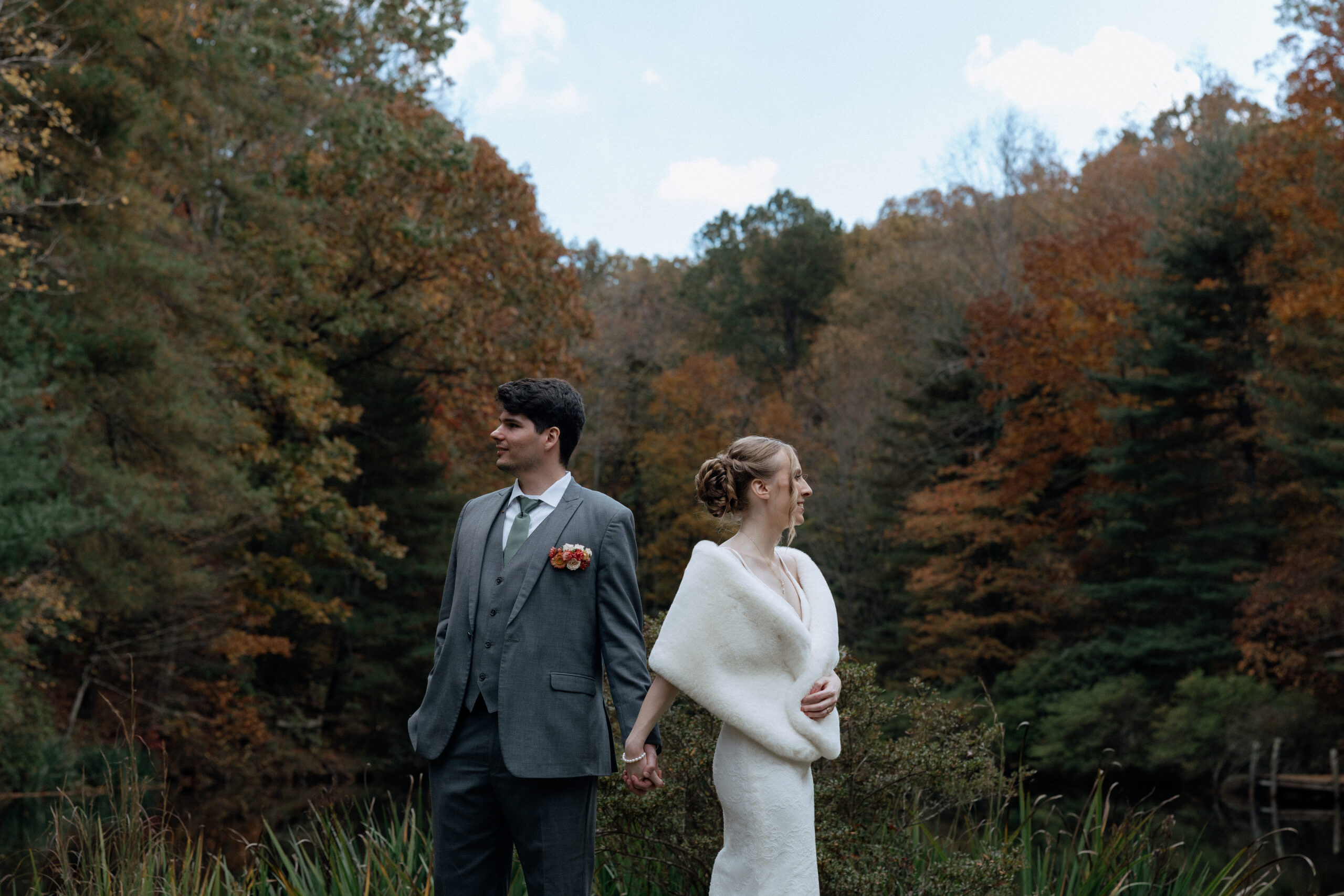 A husband and wife hold hands and look away from each other for a moody photograph in front of the autumn colored trees at Song Hill Reserve in Landrum, South Carolina.