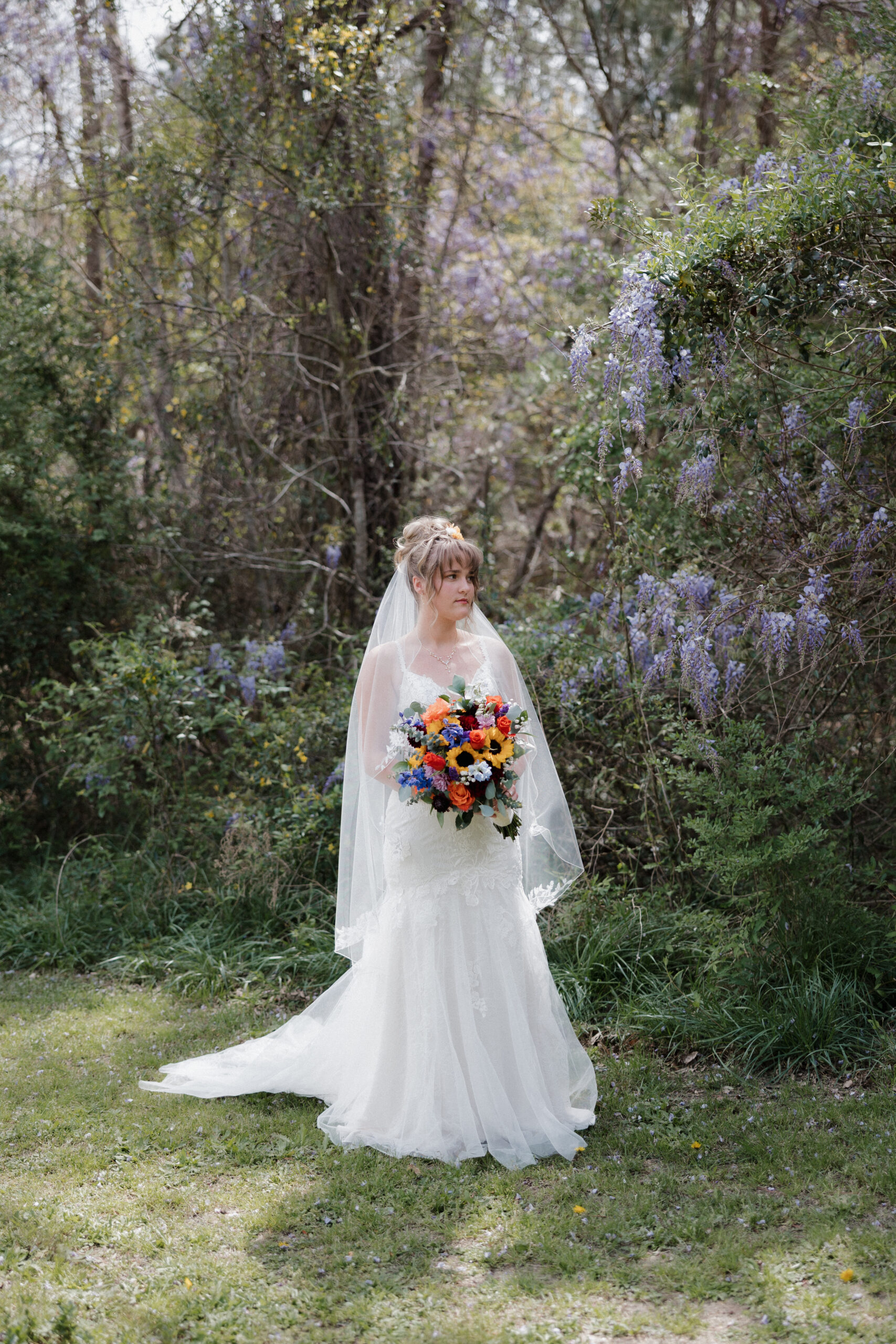 A bride holds her colorful bouquet and poses for a portrait in front of beautiful wisteria cascading from the trees in the South Carolina low country.