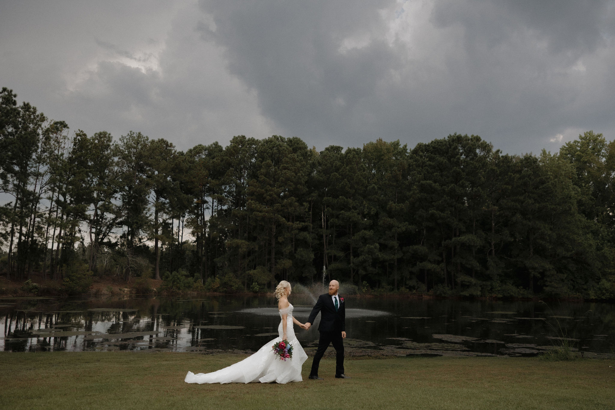 A couple holds hands and walks in front of the pond at Heron Hill in Gilbert, South Carolina on their wedding day.