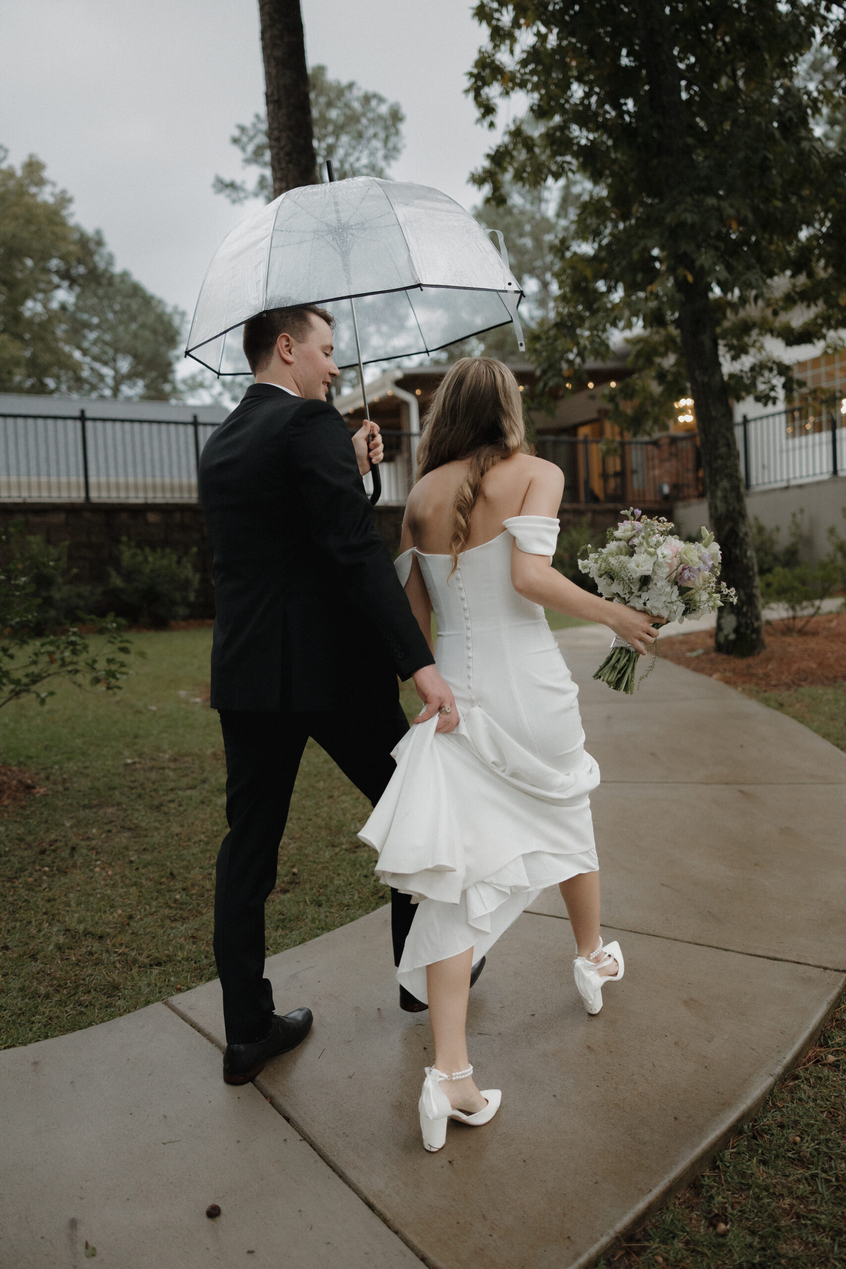 A bride and groom walk down a pathway under a clear umbrella on their rainy wedding day at Heron Hill Venue in Gilbert, South Carolina.