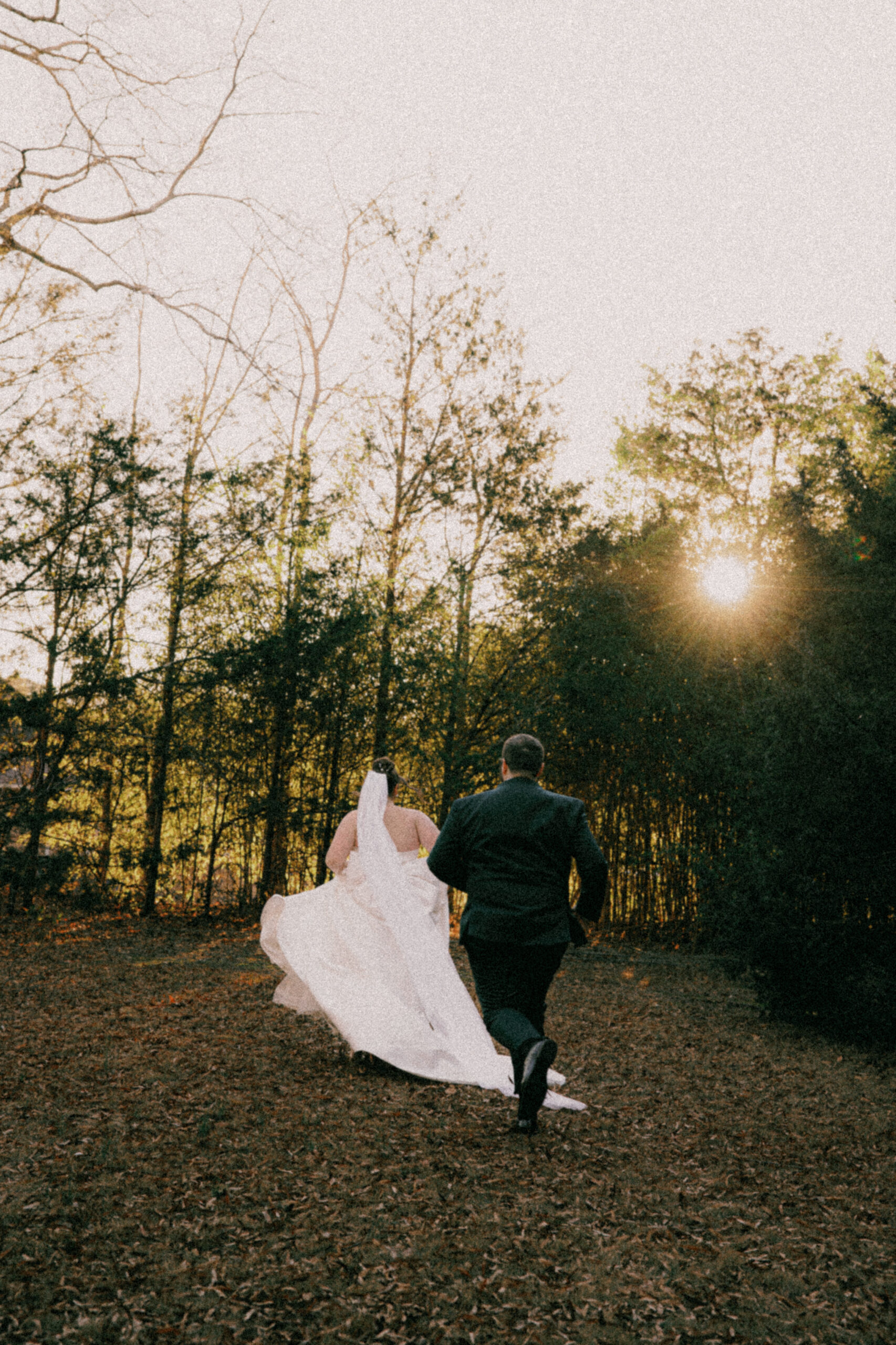 A groom runs after his bride in a field surrounded by greenery and tall trees during golden hour on their South Carolina wedding day.
